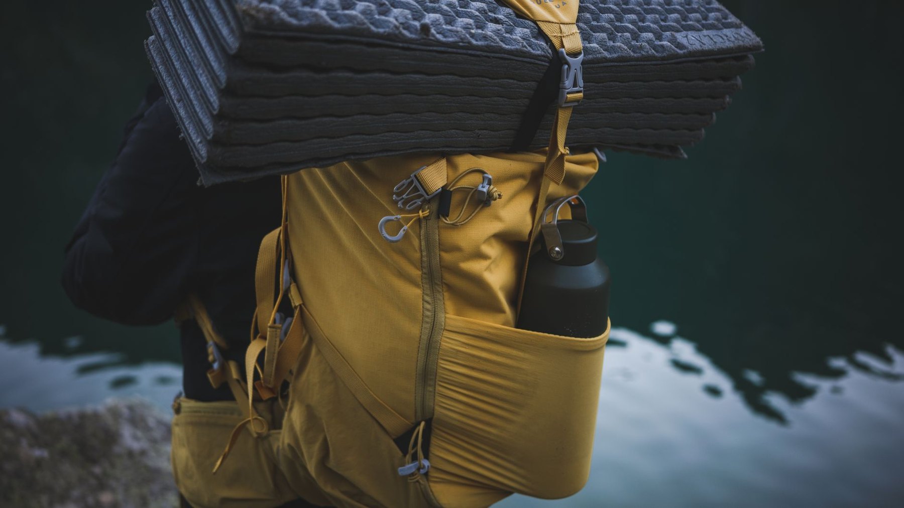 Yellow hiking backpack with a black water bottle in a side pocket and a folded foam sleeping mat strapped across the top beside water.