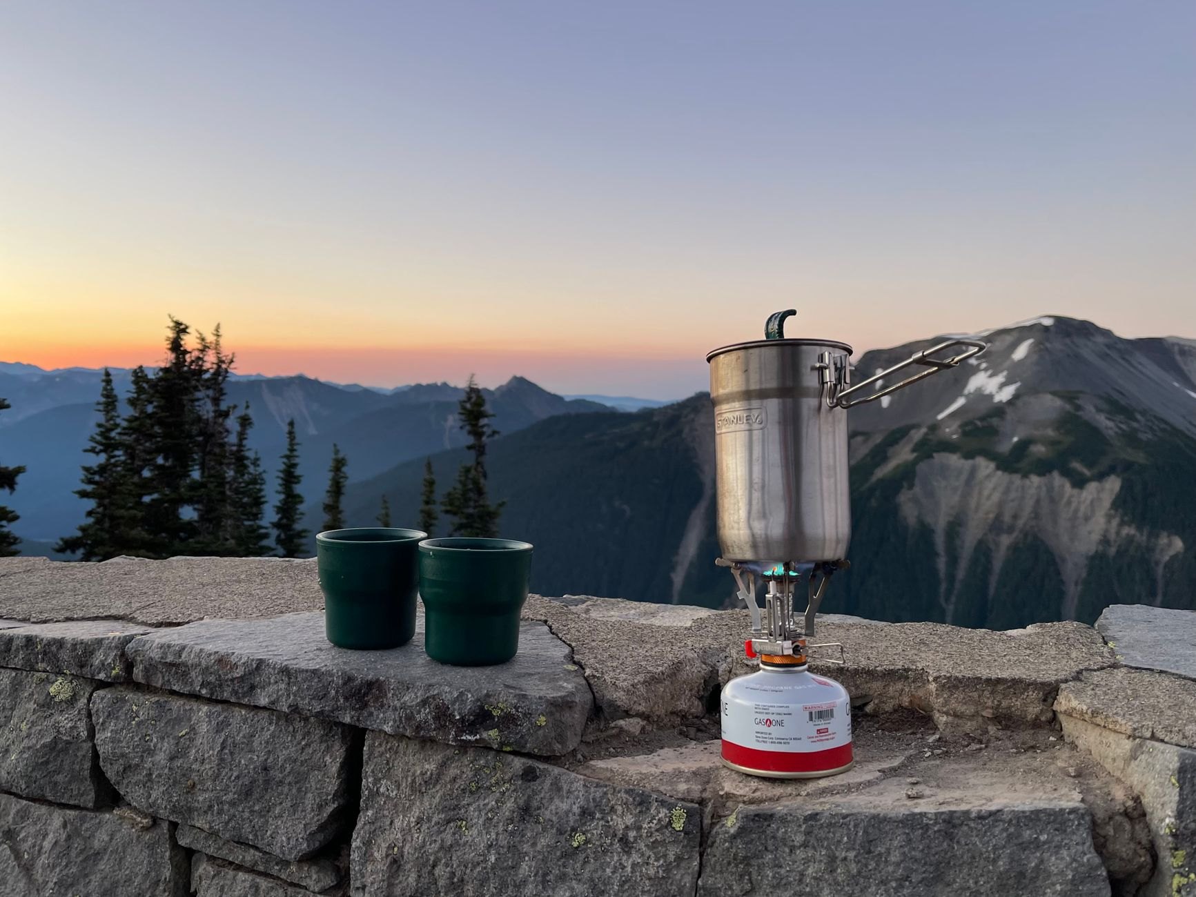A compact canister stove with a tall metal pot and two green cups on a stone wall overlooking mountains at sunrise.