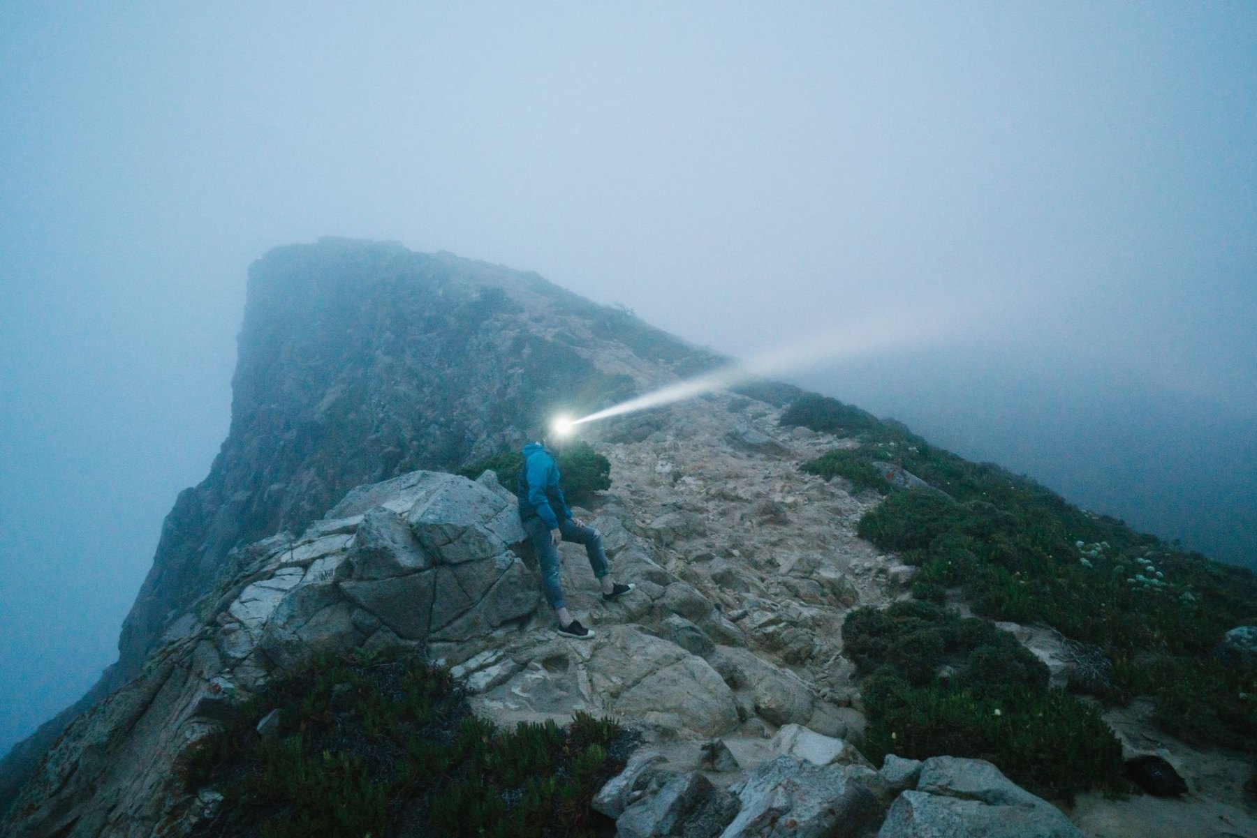 A hiker on a rocky mountain ridge in fog, with a headlamp casting a bright beam ahead.