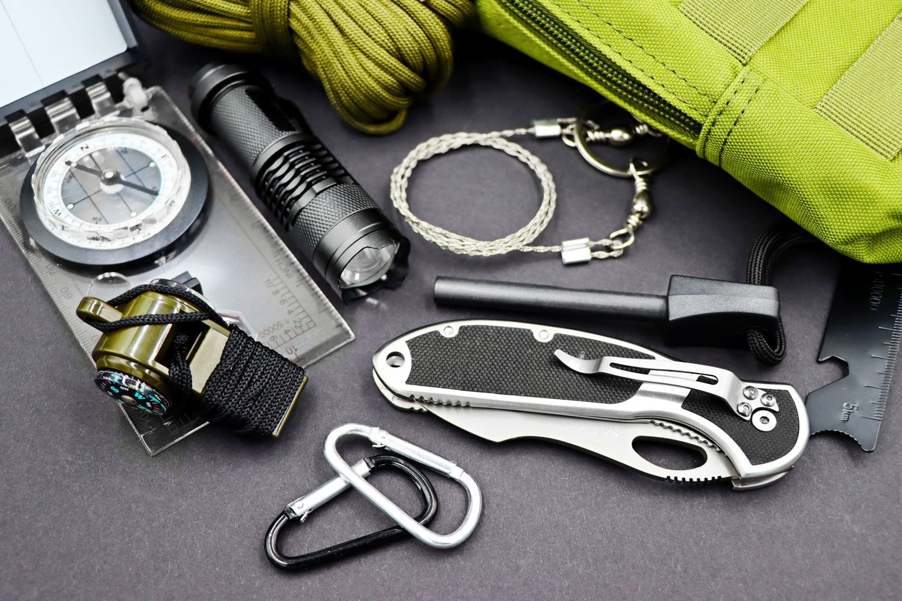 Flat lay of a compass, flashlight, folding knife, cord, wire saw, fire starter, carabiners, and green pouch on a dark surface.