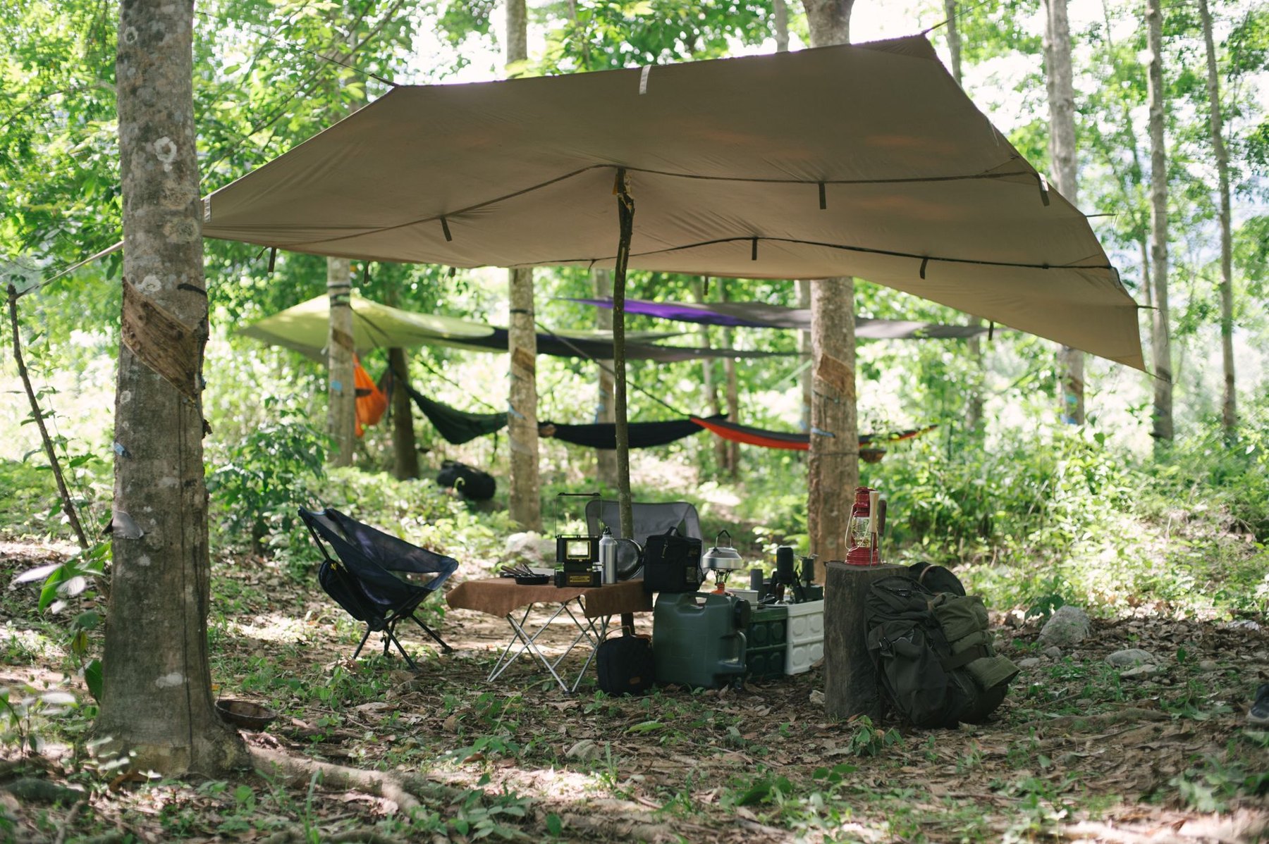 Forest campsite with hammocks strung between trees, a large overhead tarp, folding chairs, and camping gear.