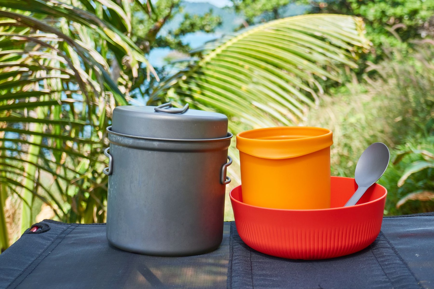 A lidded gray camping pot beside a red bowl holding an orange cup and gray spoon on an outdoor table.