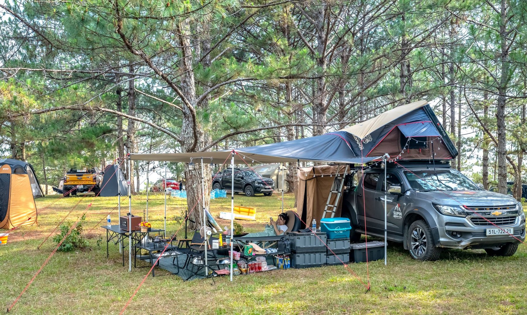 A pickup truck with a rooftop tent and extended awning beside camping tables, chairs, and storage boxes in a wooded campground.