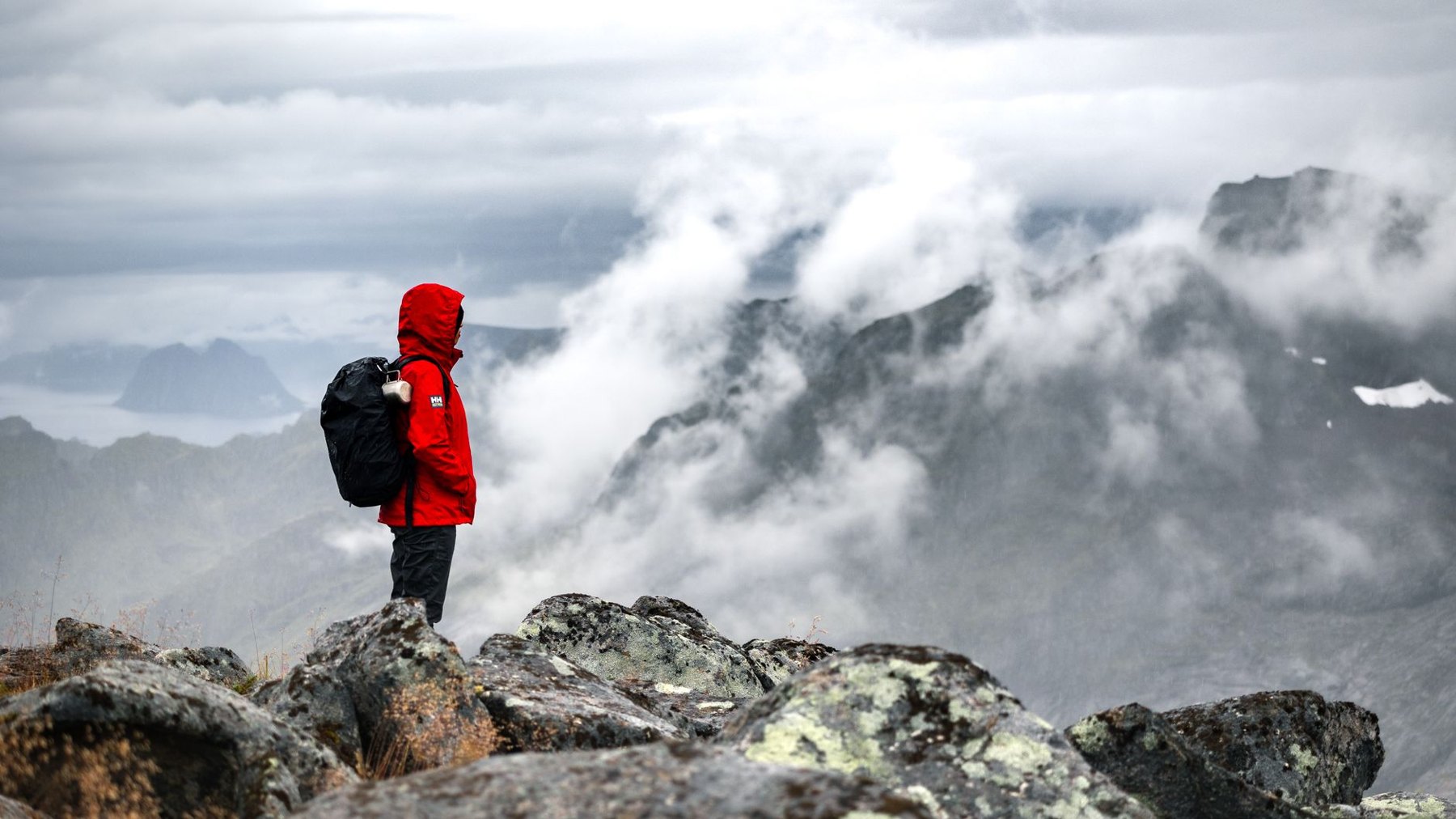A hiker wearing a red hooded jacket and a black backpack stands on rocky terrain overlooking mist-covered mountains.