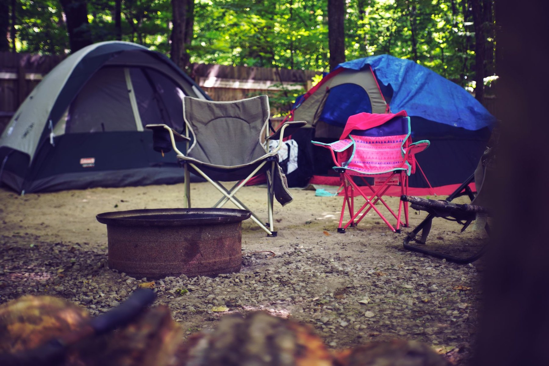 Two tents, folding camping chairs, and a metal fire ring at a wooded campsite.