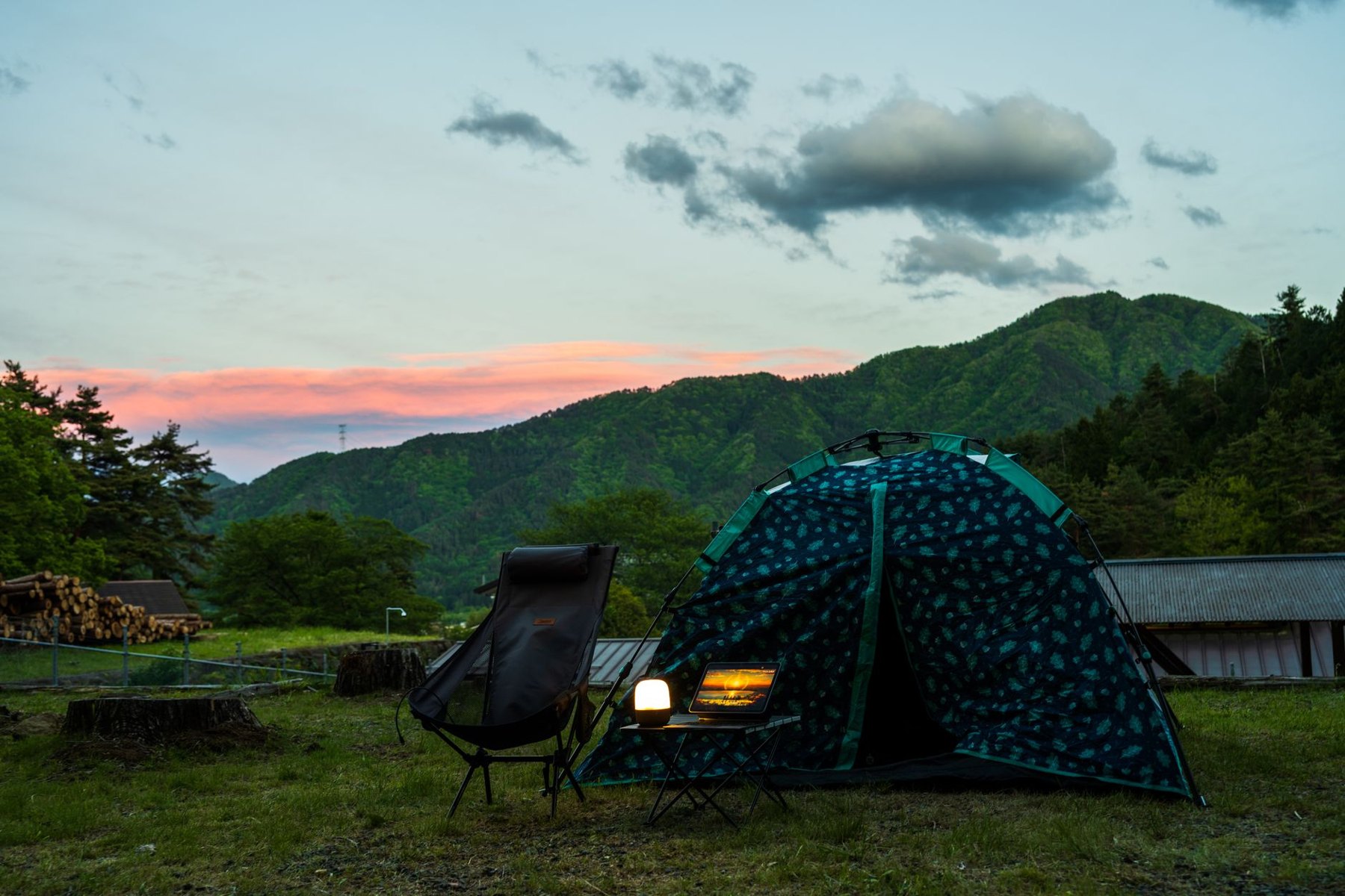 A glowing lantern beside a camping chair and tent, with wooded mountains beneath a sunset sky.