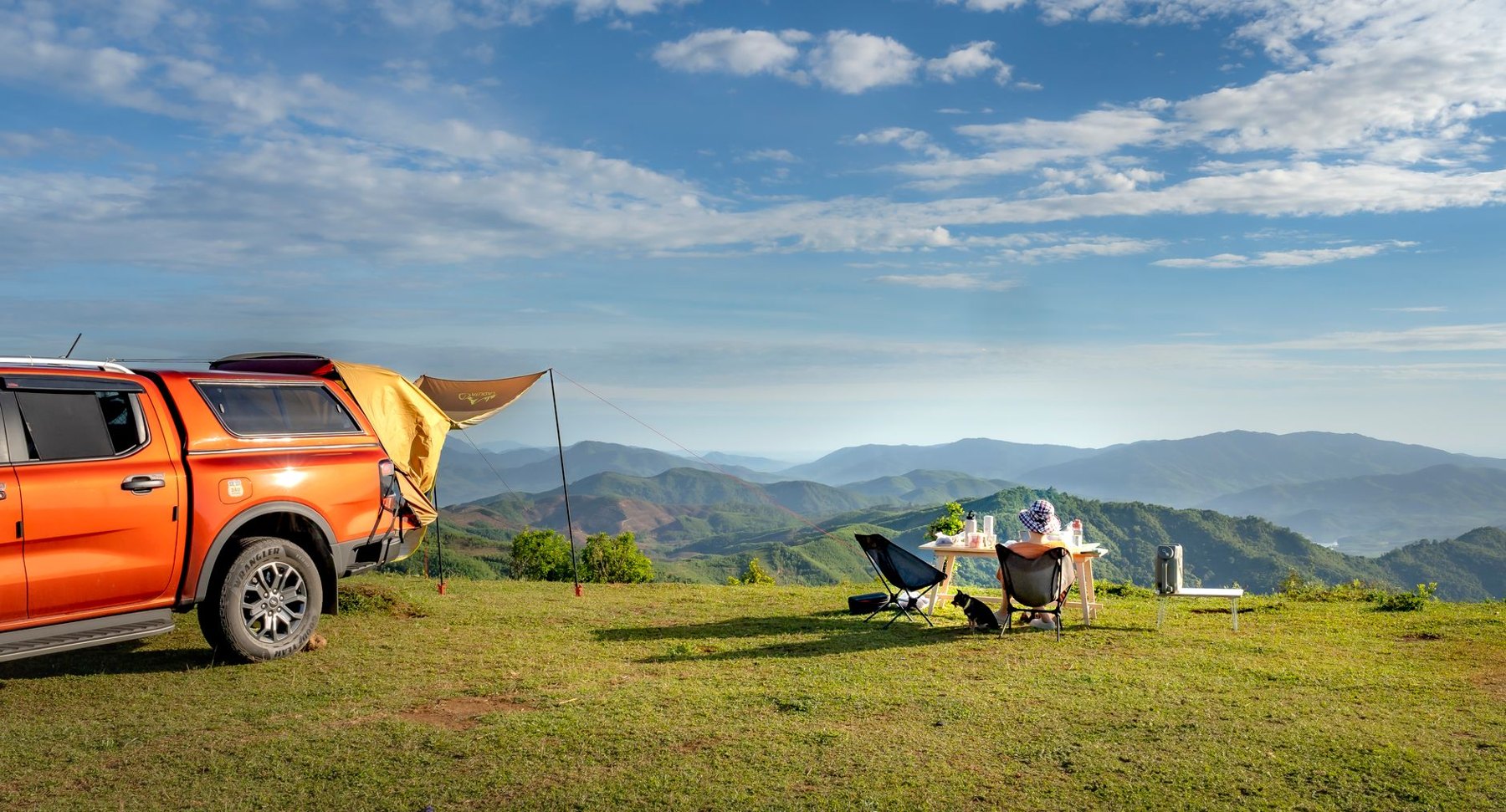 Camping chairs and a table beside an orange pickup truck at a grassy campsite overlooking mountains.