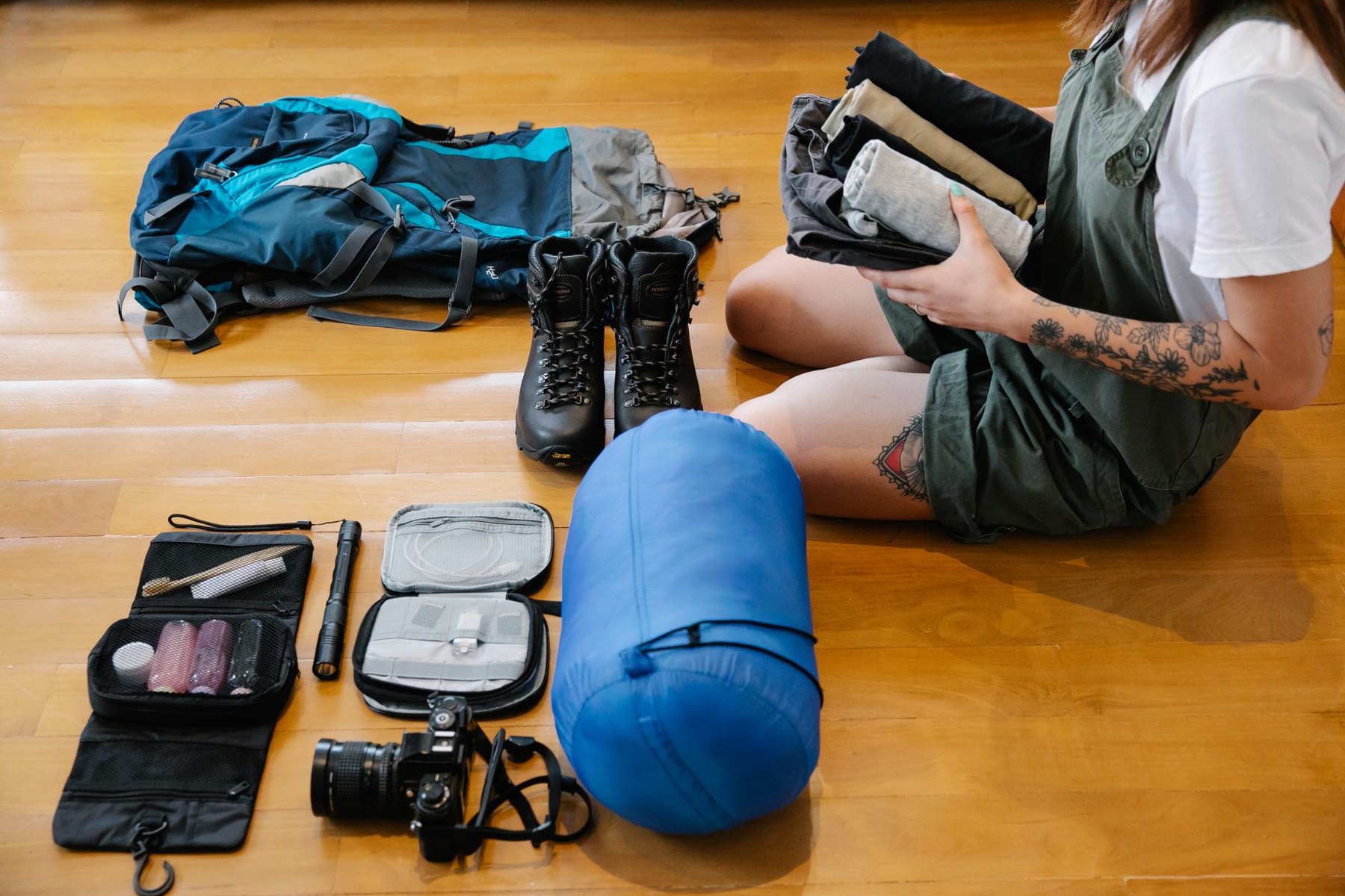 A person sorts folded clothing beside a backpack, hiking boots, a blue stuff sack, a camera, and open organizers on a wooden floor.